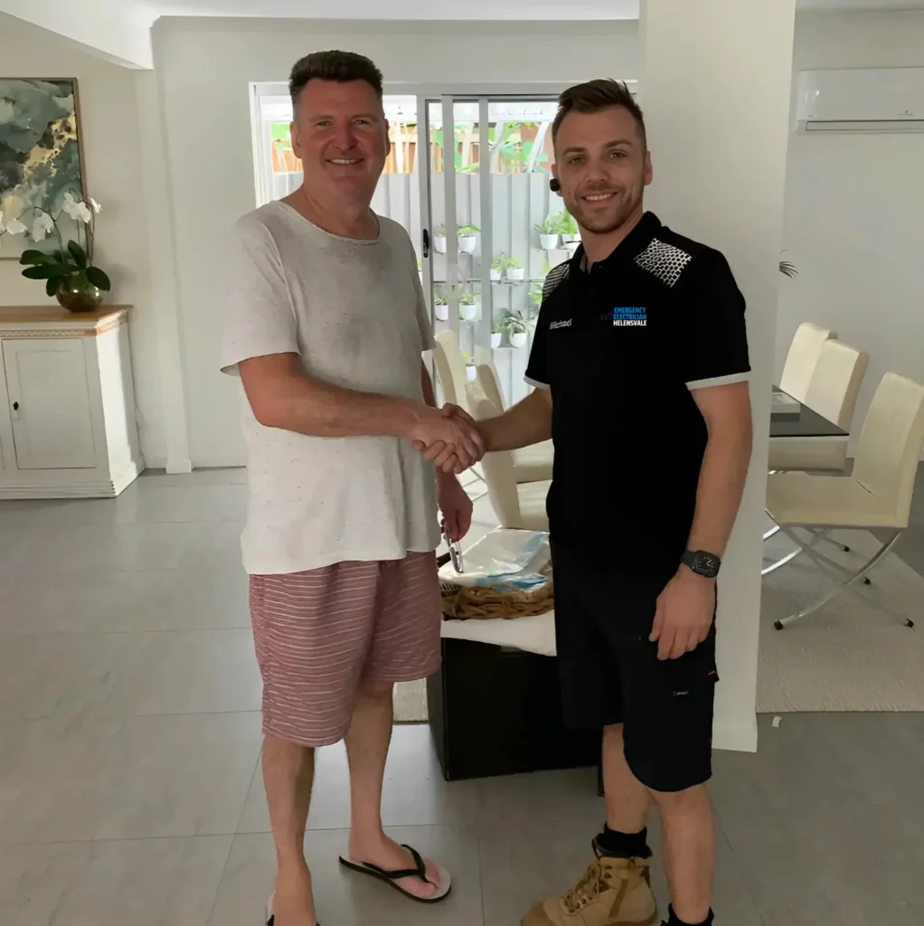 Two men shaking hands in a modern living room. One is wearing a white t-shirt and striped shorts, and the other is in a black polo shirt